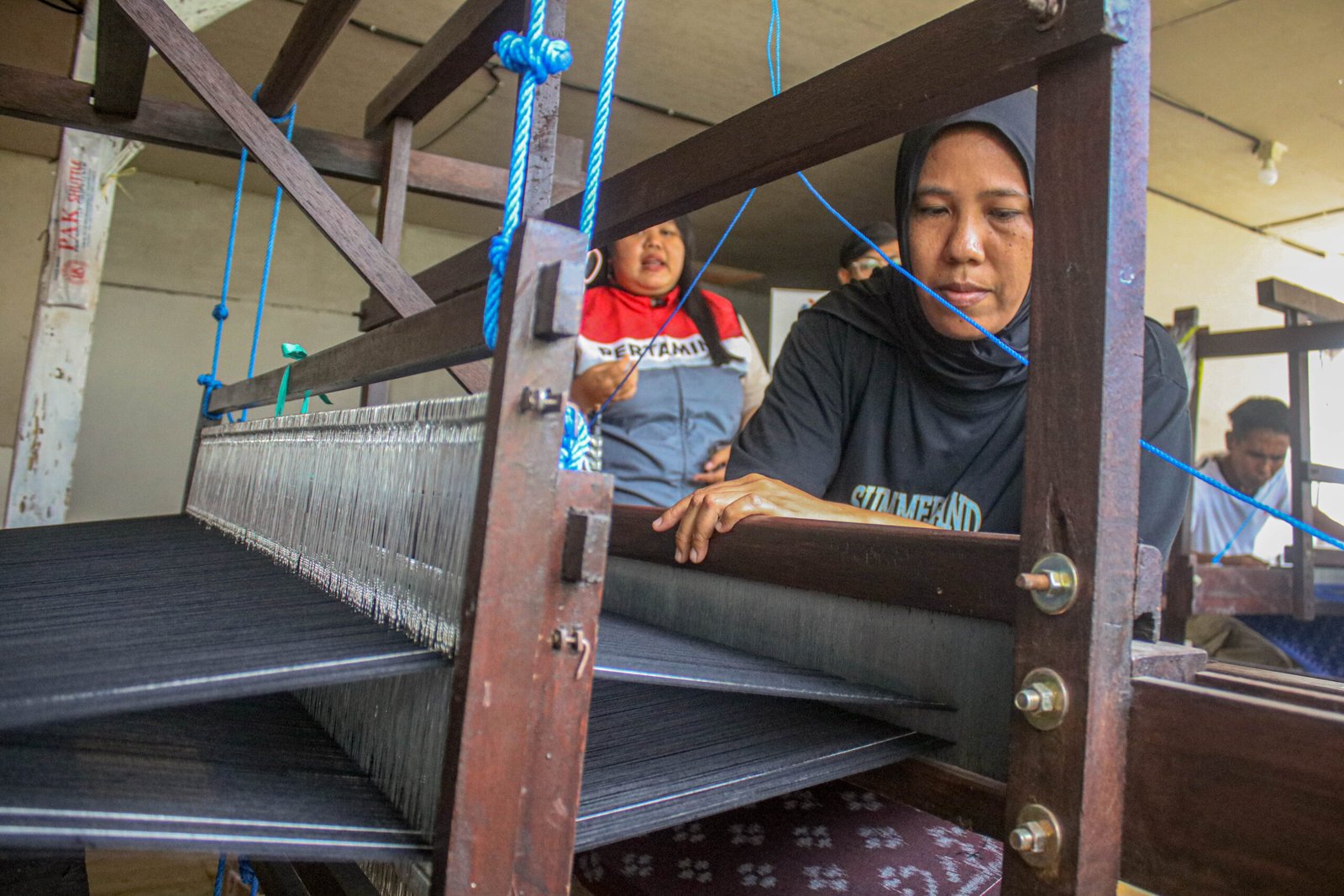 Seorang perempuan sedang melakukan proses menenun kain secara tradisional menggunakan alat tenun bukan mesin (ATBM). Dengan teliti, ia mengatur benang-benang yang tersusun rapi untuk menghasilkan kain tenun. Aktivitas tersebut berlangsung di lingkungan perajin tenun, sementara seorang anak terlihat berada di belakangnya.